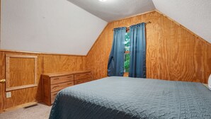 Una camera da letto con soffitto a falde, pannelli in legno, un letto con copriletto blu, una cassettiera in legno e una finestra con tende blu.