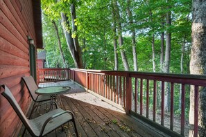 A wooden deck with two metal chairs and a small table, surrounded by a red railing, overlooking a forested area.
