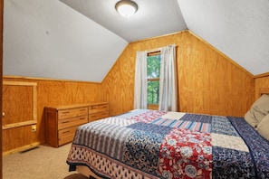 A bedroom with a wooden ceiling, a window with curtains, a bed with a quilt, and a wooden dresser.