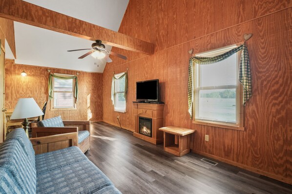 A cozy room with wooden walls, a ceiling fan, a fireplace, a television, and a sofa.