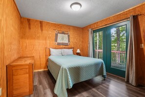 Une chambre avec un plancher en bois, un lit recouvert d’une courtepointe, une table de chevet, une fenêtre donnant sur des arbres et une porte avec un rideau.