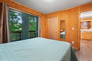 A bedroom with a large bed, wooden walls, a sliding glass door to a balcony, and a small bathroom with a mirror.