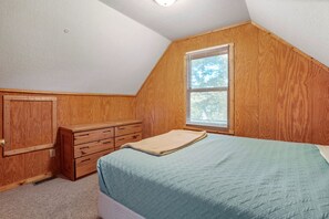 A bedroom with a sloped ceiling, wooden paneling, a window with blinds, a bed with a quilt, and a dresser with drawers. - Retreat Cabin #3 | Hocking Hills, Loft Cabin (Logan)