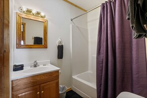 A bathroom with a wooden vanity, a mirror with lights, a bathtub, and a purple shower curtain. - Retreat Cabin #3 | Hocking Hills, Loft Cabin (Logan)