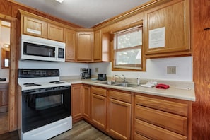 A kitchen with wooden cabinets, a microwave, stove, sink, and a window with blinds. - Retreat Cabin #3 | Hocking Hills, Loft Cabin (Logan)