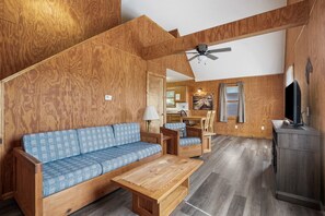 A wooden-floored living area with a sofa, chairs, a coffee table, and a TV stand. - Retreat Cabin #3 | Hocking Hills, Loft Cabin (Logan)