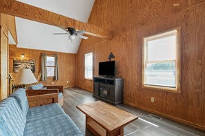 A spacious living room with a wooden ceiling, a ceiling fan, a television on a cabinet, a sofa, a coffee table, and large windows. - Retreat Cabin #3 | Hocking Hills, Loft Cabin (Logan)