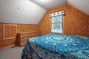 Ein Schlafzimmer mit Holzboden, einem Fenster mit Jalousien, einem Bett mit gemustertem Überwurf und einer Holzkommode.