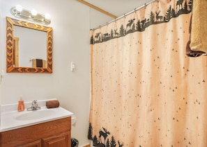 Une salle de bain avec un meuble-lavabo en bois, un miroir avec éclairage intégré, un rideau de douche orné d’un motif de forêt et une serviette accrochée au mur.