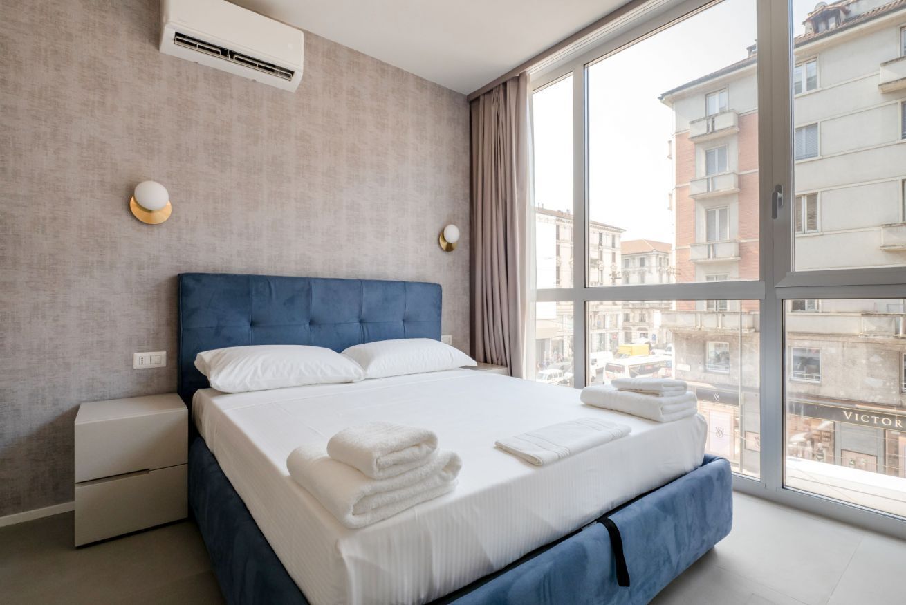 A modern hotel room with a large bed, a nightstand, an air conditioning unit, and a view of the city through large windows.