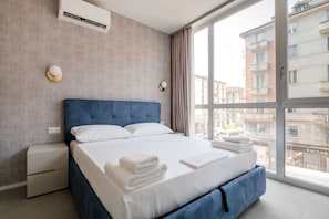 A modern hotel room with a large bed, a nightstand, an air conditioning unit, and a view of the city through large windows.