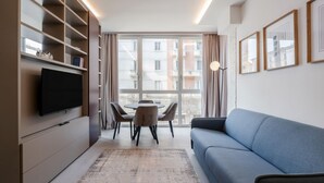 A modern living room with a flat-screen TV, a blue sofa, a dining table with chairs, and a large window with curtains. - GuestHost - Porta Venezia - Modern Studio near Corso Buenos! (Milano)