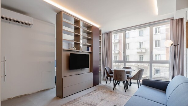 A modern living room with a flat-screen TV, built-in shelving, a sofa, a dining table with chairs, and large windows. - GuestHost - Porta Venezia - Modern Studio near Corso Buenos! (Milano)