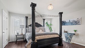 Ein Bett mit Baldachin, dunkel gehalten, ein Nachttisch mit einer Vase und ein an der Wand befestigtes Regal mit einer Pflanze.