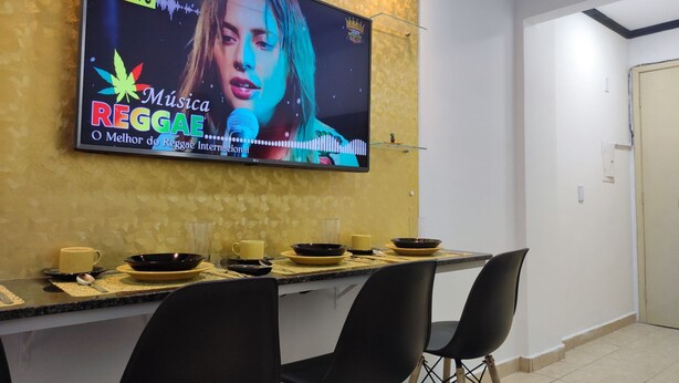 A dining area with a TV displaying a music event, a table set with bowls and cups, and two chairs. - Renomado Apartamento em Curitiba  (Curitiba)