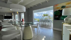 Un moderno salón con una mesa de comedor de vidrio, muebles blancos y un amplio ventanal con vistas al océano.