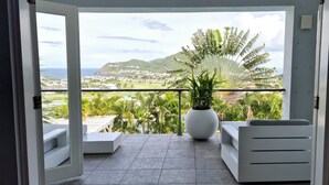 Un balcón con una silla blanca, una planta grande en maceta y vistas al paisaje costero.