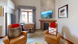 Un salón con dos sillones de cuero, una mesa de comedor de madera, una chimenea, un televisor de pantalla plana montado en la pared y un ventanal con cortinas.