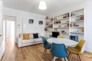 A modern living room with a white sofa, a round table, and colorful chairs. There are bookshelves filled with books and a television on a stand.