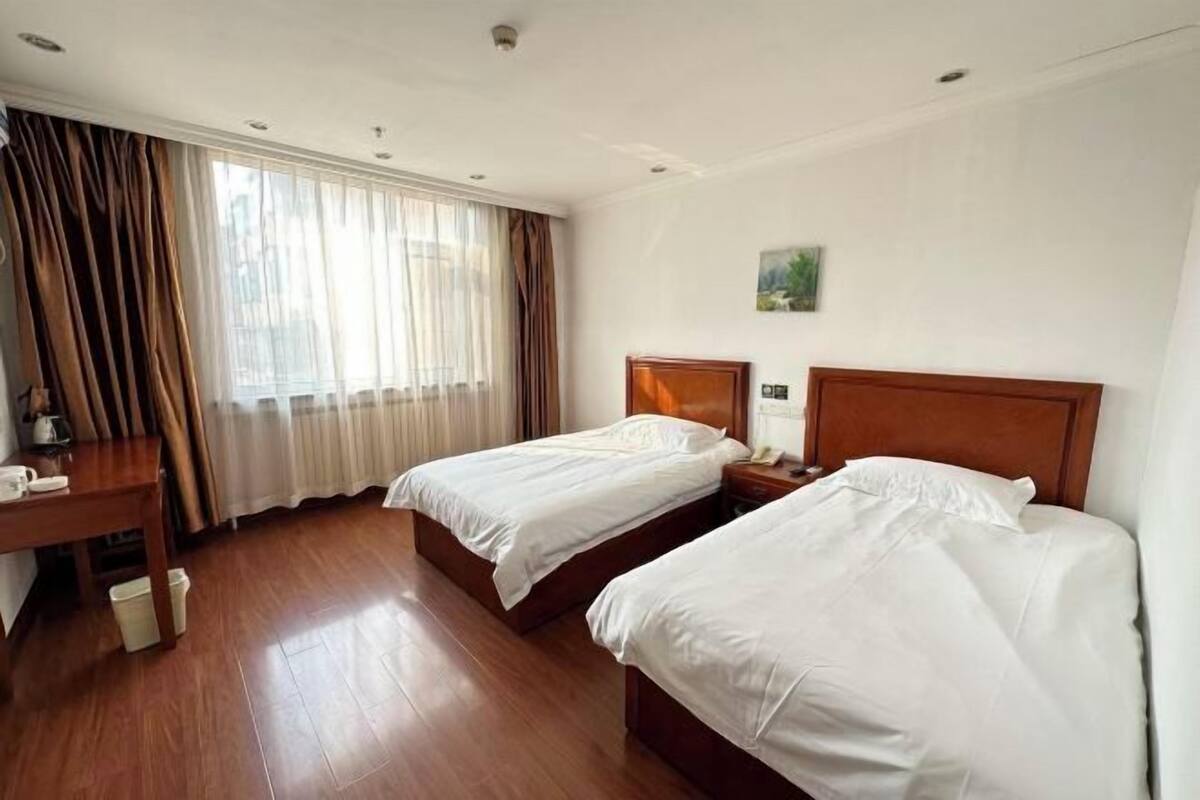 A hotel room with two beds, a wooden floor, a desk with a chair, a window with curtains, and a painting on the wall.