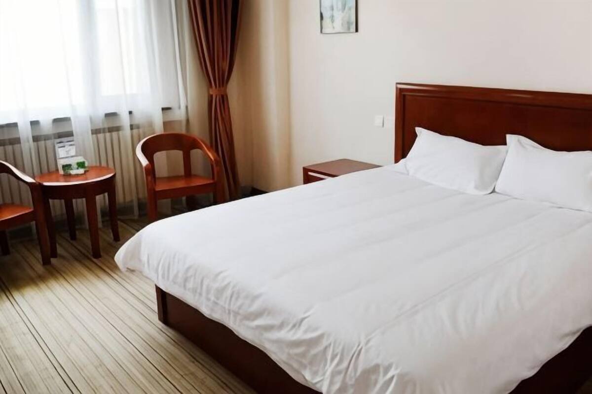 A hotel room with a wooden bed, a small bedside table, a chair, and a window with curtains.