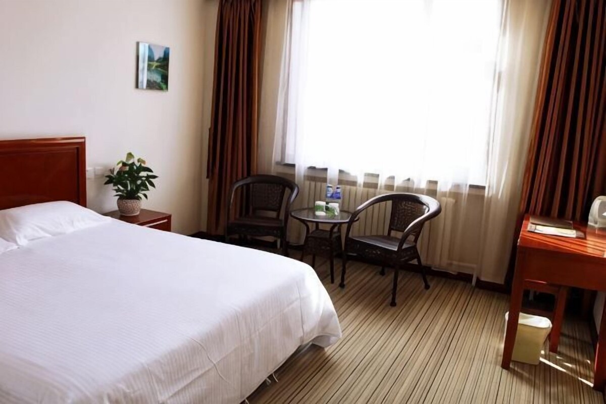 A hotel room with a bed, a small table with a plant, two chairs, a desk, and a window with curtains.