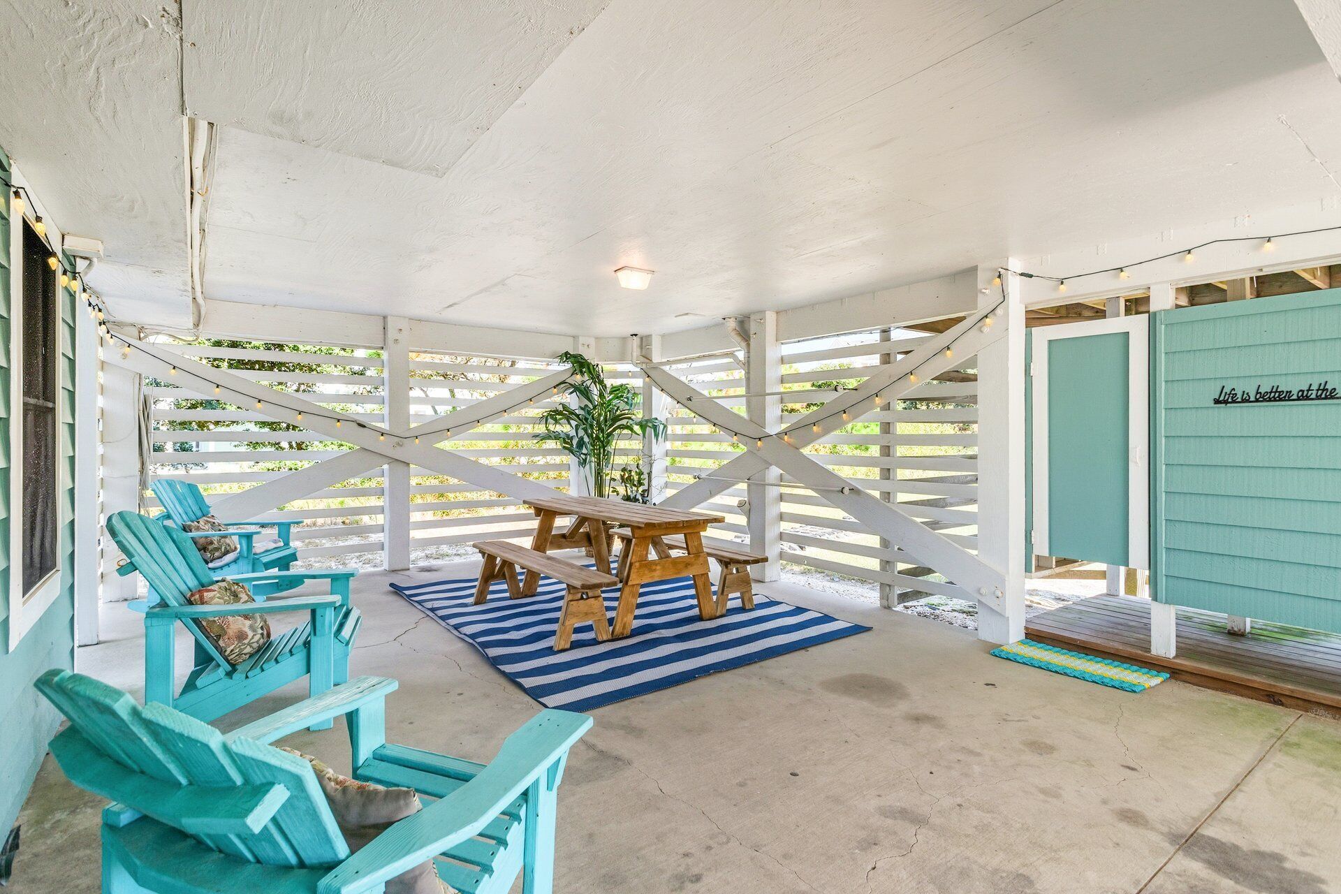 Un espace extérieur aménagé avec des meubles en bois et des chaises turquoise, protégé par un auvent, et agrémenté d’un tapis à rayures et de guirlandes lumineuses.