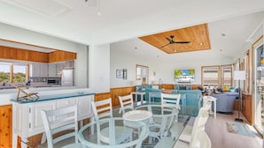 A spacious living area with a glass dining table, white chairs, a blue sofa, and a wooden ceiling. - Dolphin Days Hot Tub, Pool & steps to the Beach (Corolla)