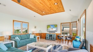 A spacious living room with a wooden ceiling, a television mounted on the wall, and a view of the beach through large windows. - Dolphin Days Hot Tub, Pool & steps to the Beach (Corolla)