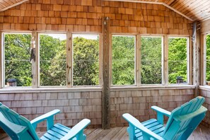 Une véranda couverte aux murs en bois et aux grandes fenêtres, deux fauteuils Adirondack bleus et une vue sur les arbres.