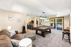 Ein Billardtisch, eine Couch, ein Barhocker und ein Essbereich mit Stühlen sowie eine Küche im Hintergrund.