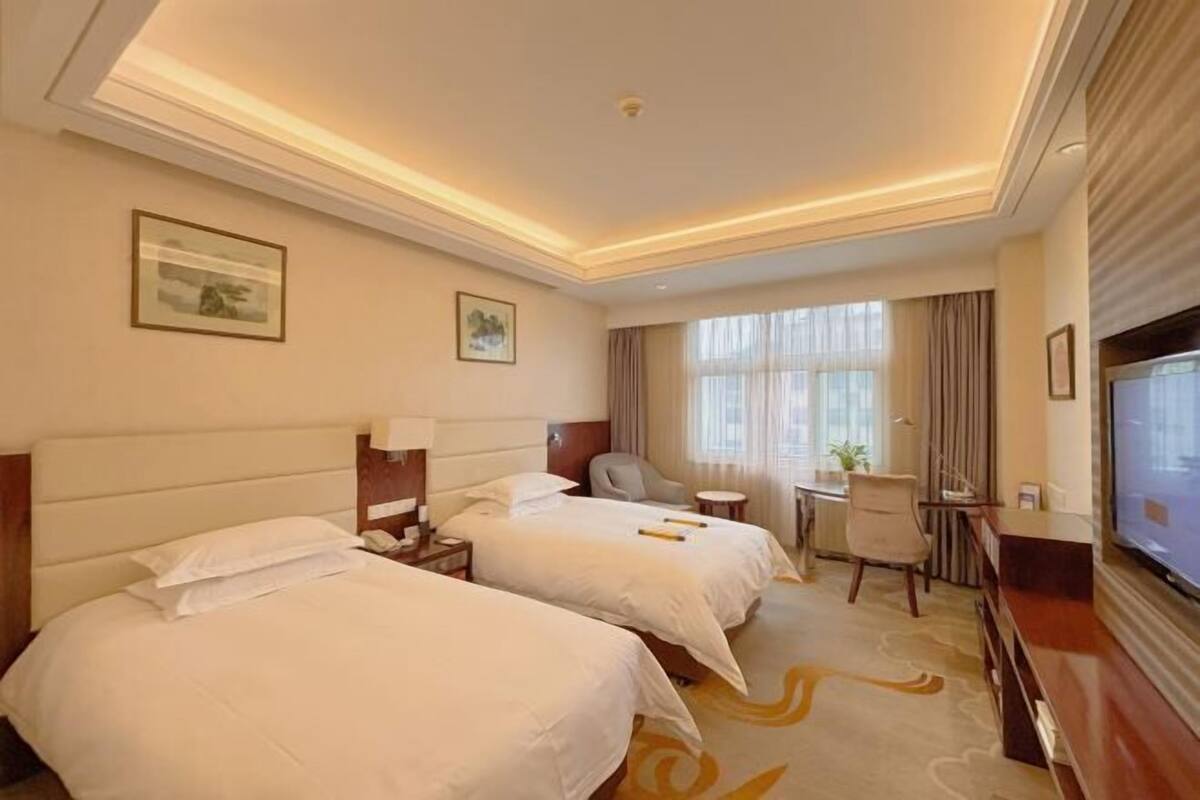 A hotel room with two beds, a desk, a chair, a TV, and a window with curtains.