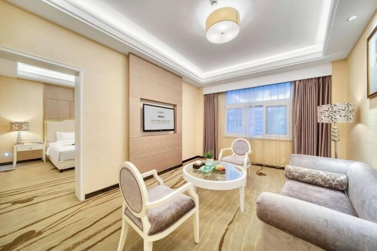A hotel room with a bed, a sitting area with a round table and chairs, a large window with curtains, and a framed certificate on the wall.