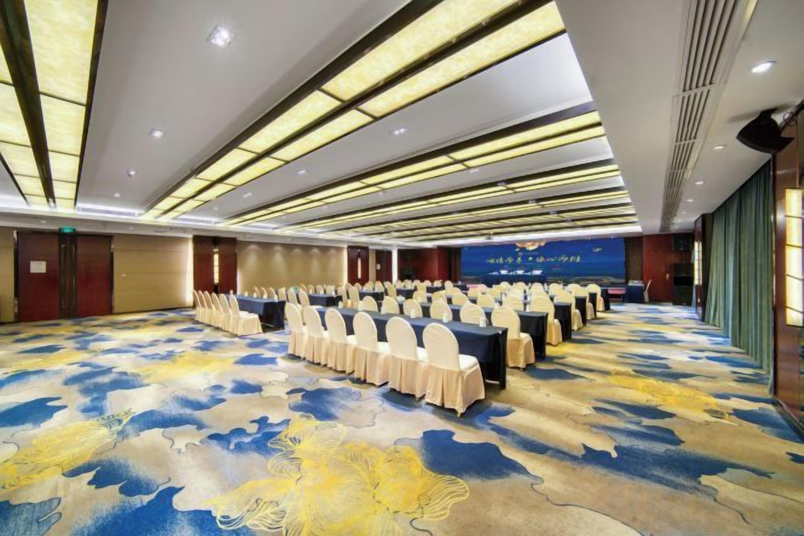 Une salle de conférence avec des rangées de tables et de chaises, une moquette à motifs et un fond bleu sur lequel est inscrit du texte.