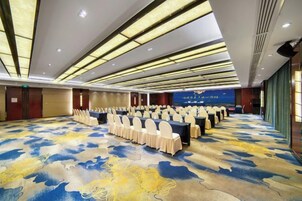 Un salón de conferencias con filas de mesas y sillas, alfombra estampada y un fondo azul con texto.