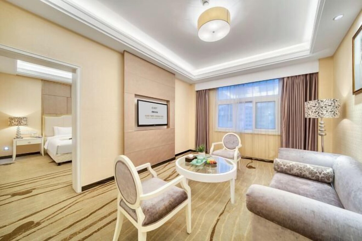 A hotel room with a bed, a sitting area with a round table, a sofa, a lamp, and a framed certificate on the wall.