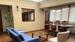 Ένα σαλόνι με έναν καναπέ, μια πολυθρόνα, έν α τραπέζι τραπεζαρίας με καρέκλες και ένα μεγάλο παράθυρο με κουρτίνες.
