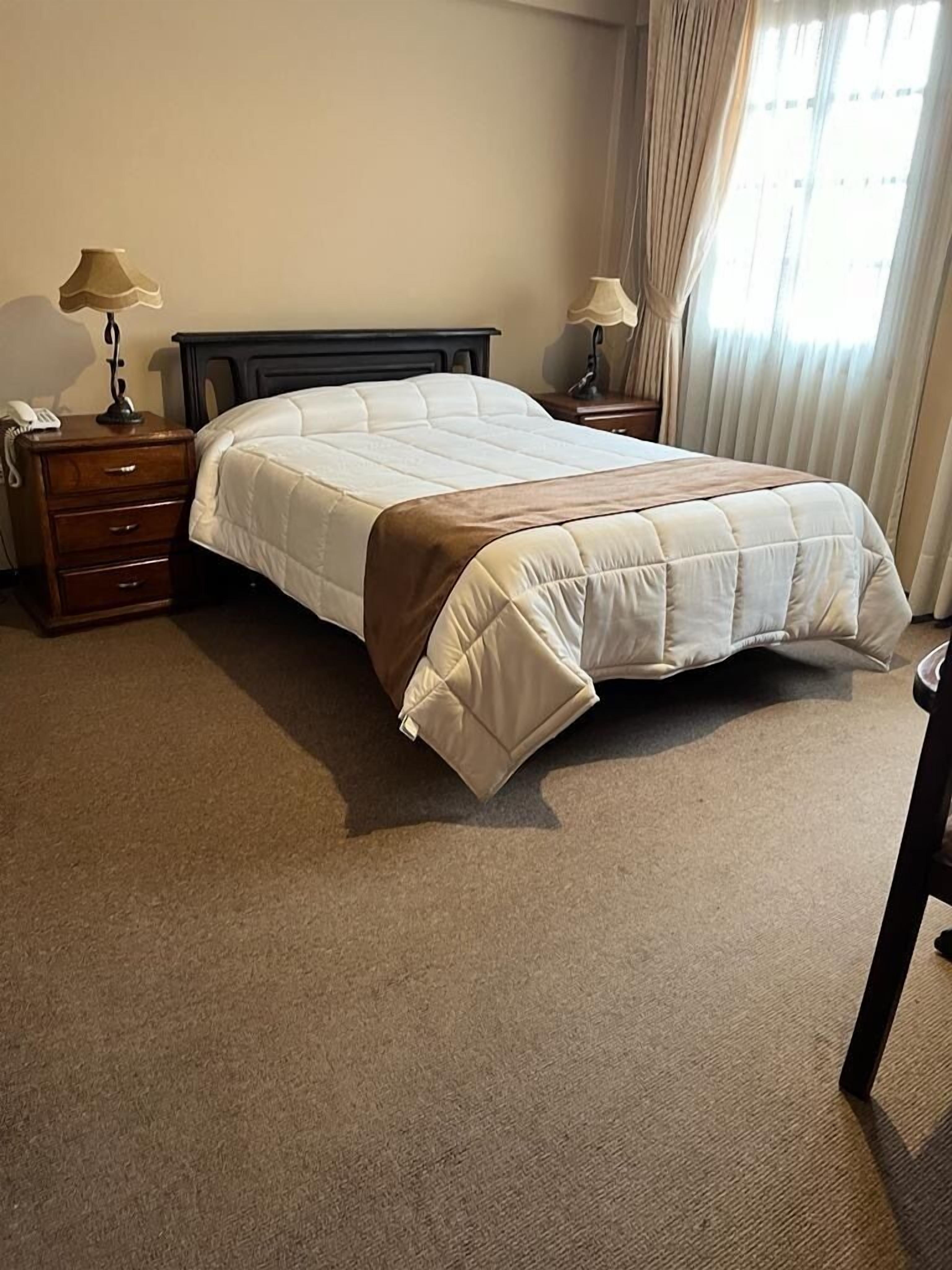 A neatly made bed with a white duvet and brown throw, flanked by wooden nightstands with lamps, in a room with beige carpet and light-colored walls.