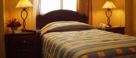 Quarto de hotel com cama, duas luminárias de cabeceira, mesa de apoio, telefone e janela com cortinas.