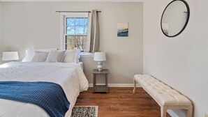 A bedroom with a bed, bedside lamps, a window with curtains, a bedside table, a bench, and a mirror on the wall. - Designer Home - Pool - Billiards - Home Office (Cary)