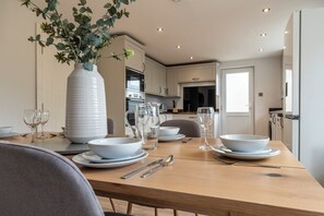 A dining table set with white plates, bowls, and wine glasses, a large white vase with green foliage, and a modern kitchen in the background. - Bright&Spacious 3BD in Centre of Bourton (Cheltenham)
