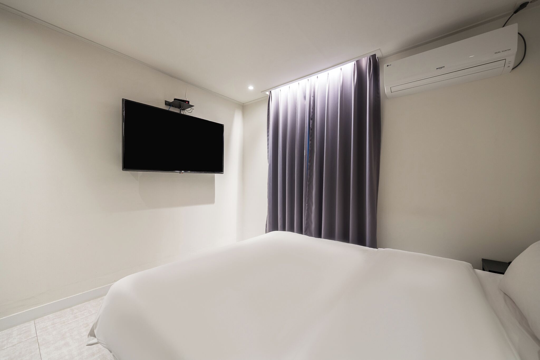 A hotel room with a bed, a flat-screen TV mounted on the wall, and an air conditioning unit.