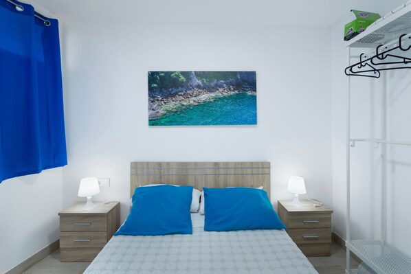 A bedroom with a bed, two bedside tables with lamps, a wall-mounted picture, and a metal rack.