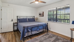A bedroom with a bed, a ceiling fan, a window with blinds, a door, and a small table with a lamp. - Little Mountain Cabin - 3-Bedroom Retreat with Hot Tub, Fire Pit, Games, and Serene Nature Views (Sevierville)