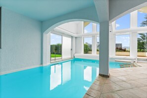A swimming pool with clear blue water, white walls, and large windows offering views of greenery and buildings.