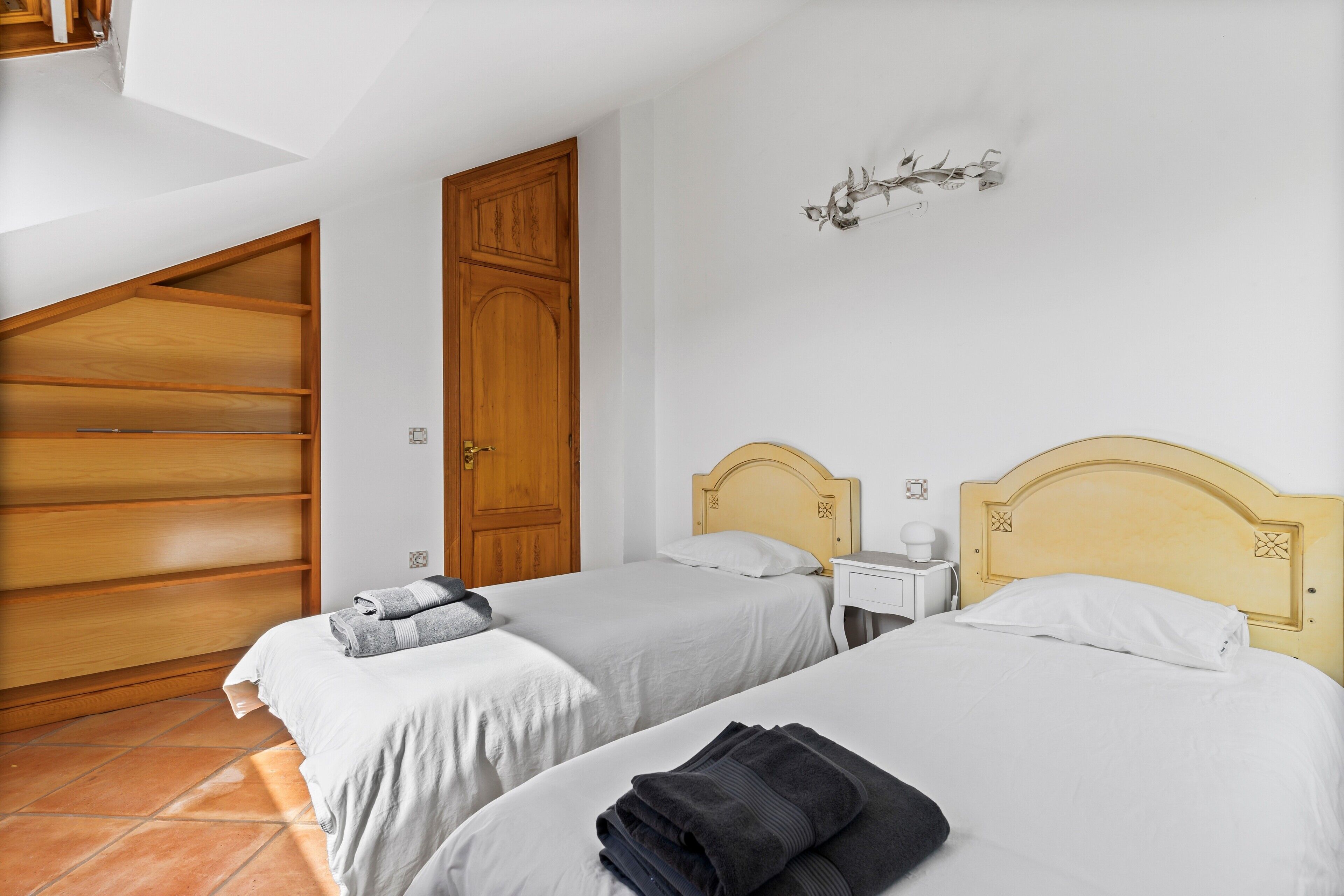 A room with two beds, a wooden wardrobe, a nightstand, and a wall-mounted light fixture.