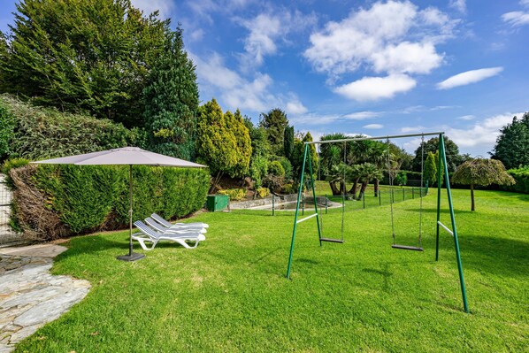 A grassy outdoor area with a swing set, a covered seating area, and a stone pathway.