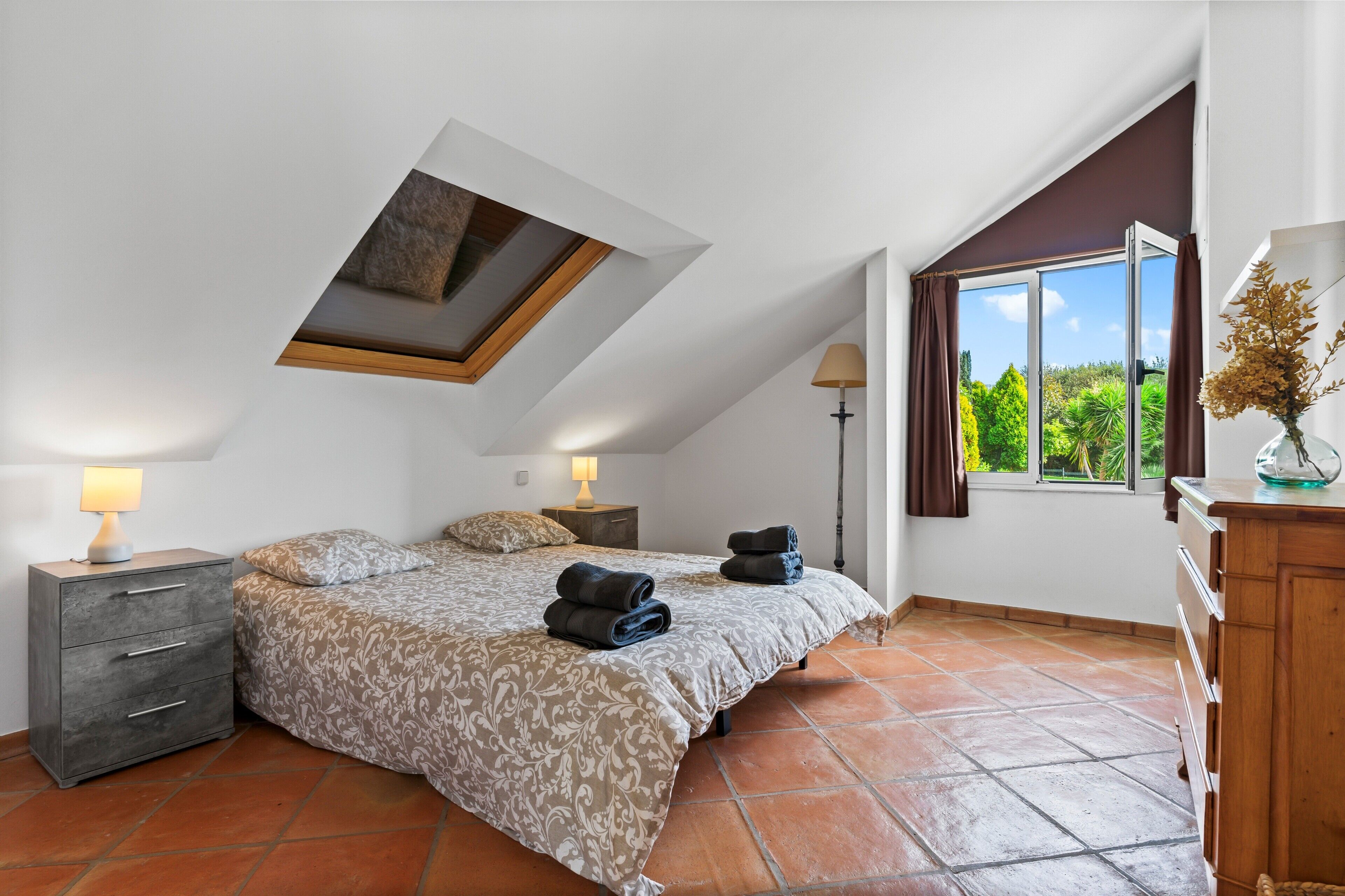 Een slaapkamer met dakraam, een bed, nachtkastje, dressoir en raam met gordijnen.