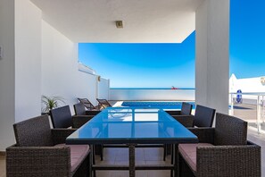 Un espace extérieur aménagé, abrité et doté d’une table en verre et de chaises en osier, offrant une vue sur une piscine et un ciel d’un bleu limpide.
