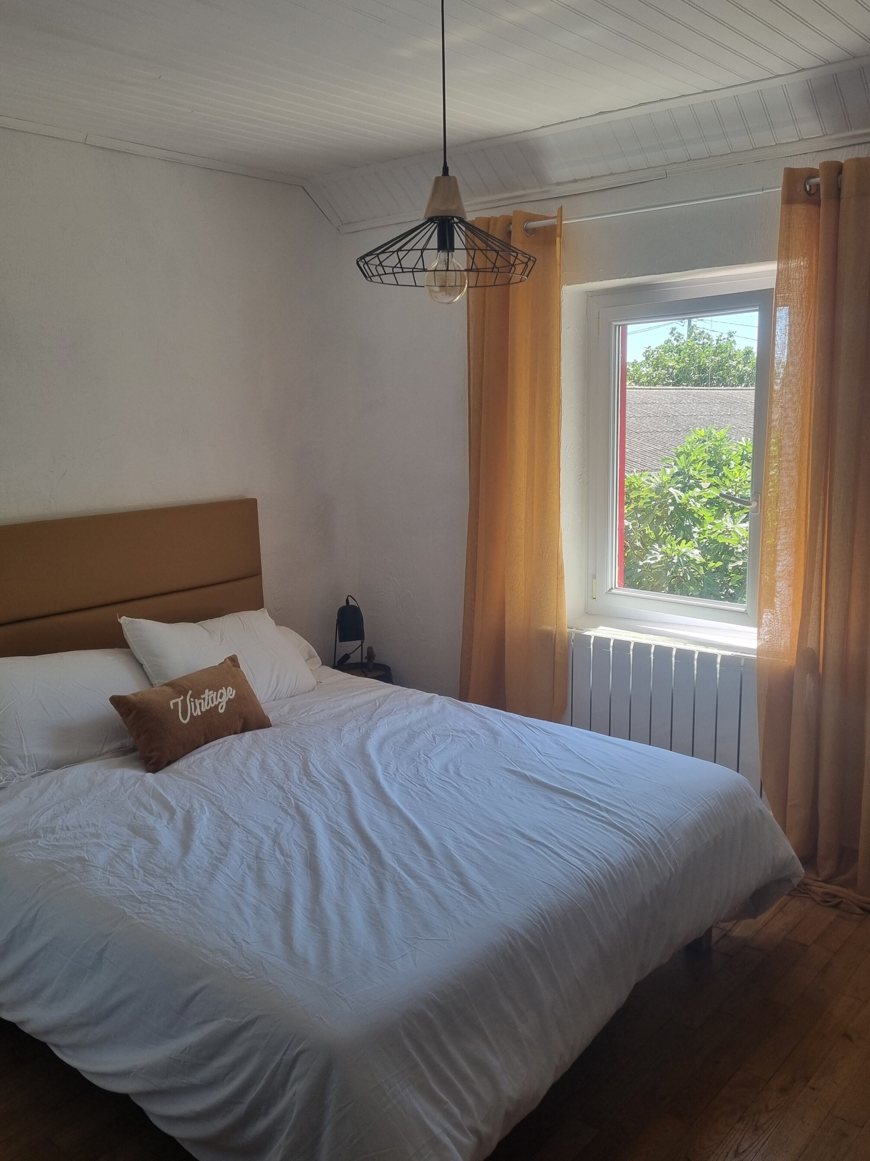 Una cama bien hecha con sábanas blancas y un almohadón marrón con la etiqueta 'Vintage', en una habitación que tiene un cabecero de madera, una mesita de noche, una lámpara y una ventana con cortinas.